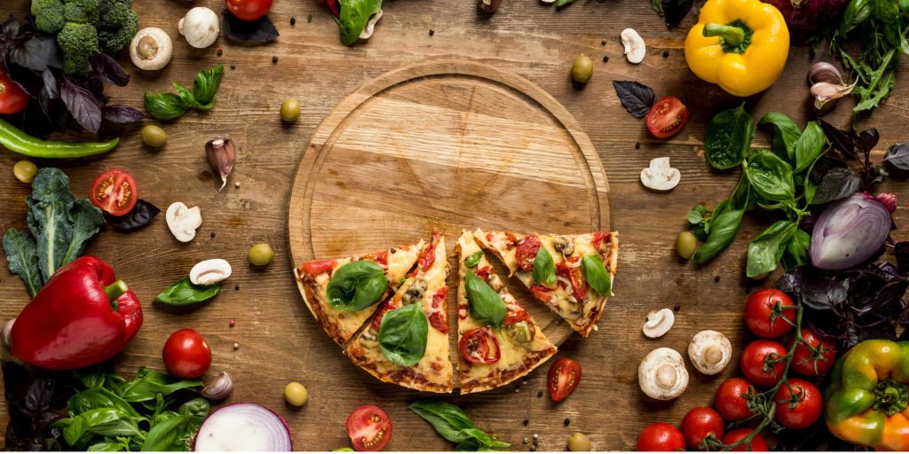 italian pizza slices on wooden board with various fresh vegetables and herbs on table